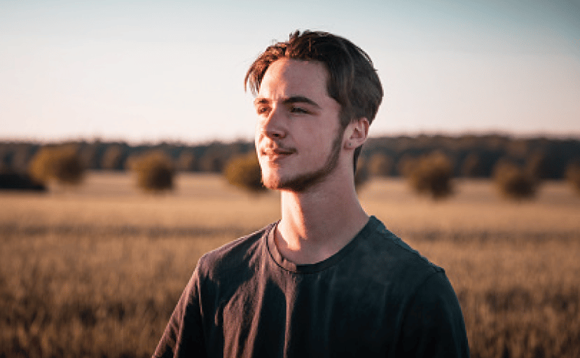 Young man in a dark shirt stands in a sunlit field, looking into the distance with trees and an open landscape in the background.