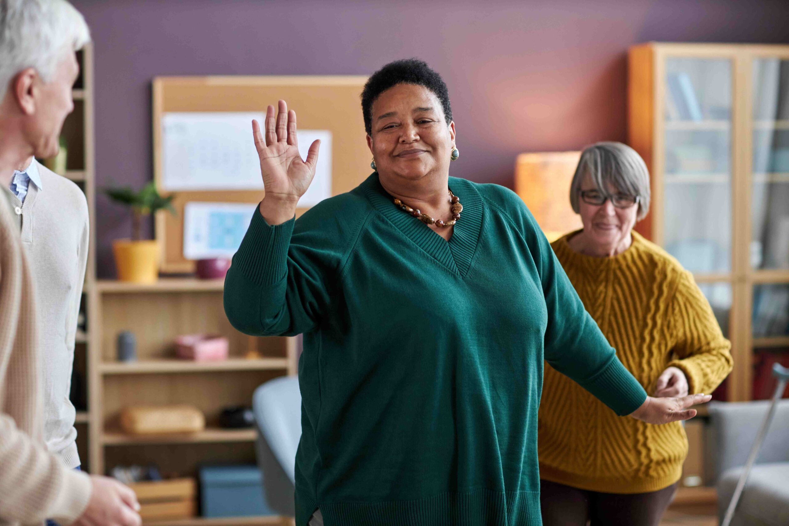 In a room with a bookshelf and a board, a person in a green sweater is smiling and raising a hand, perhaps sharing their journey through alcohol detox with two others listening intently.