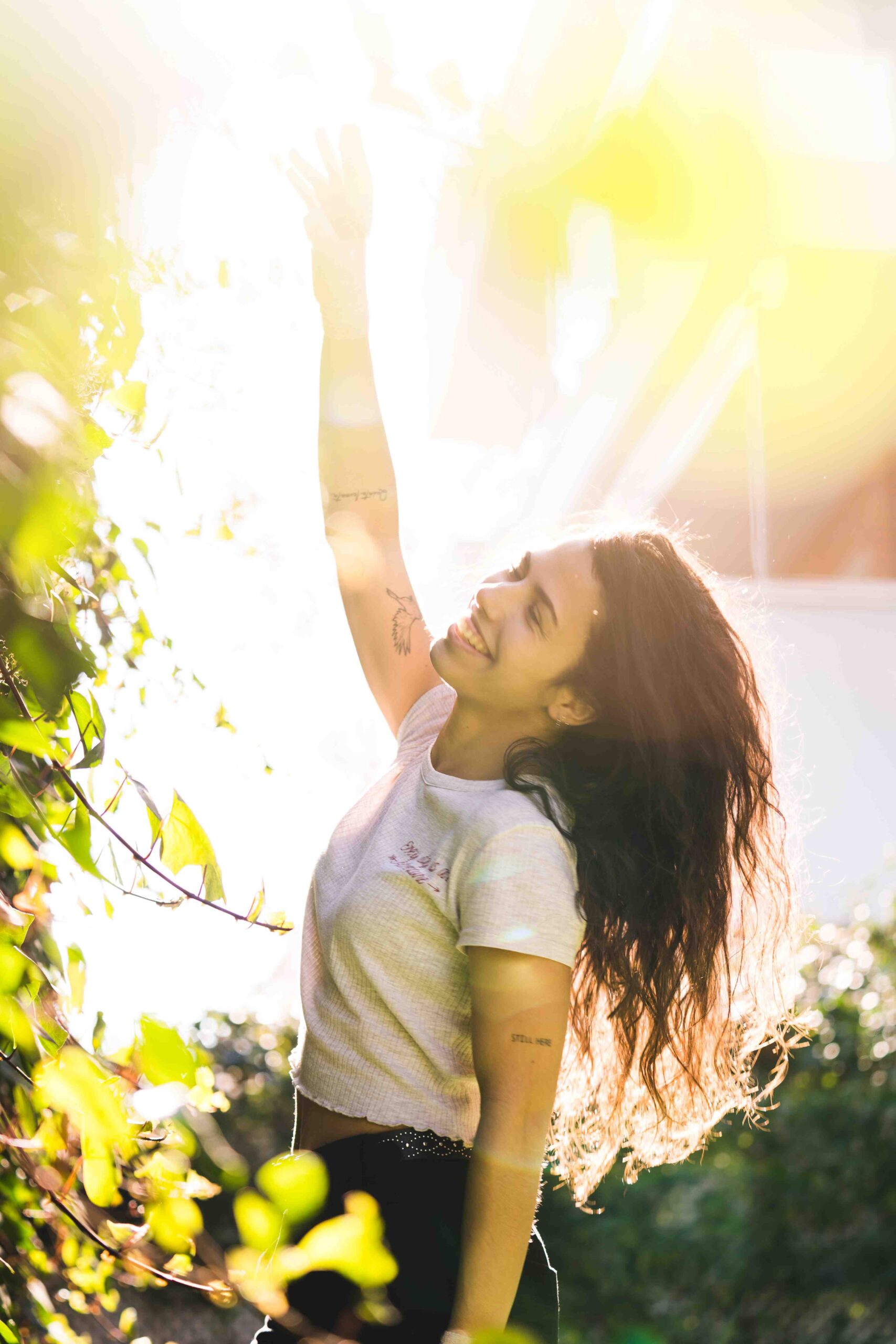 A woman with long curly hair stands outdoors, smiling and reaching towards the sun, surrounded by greenery, embracing a moment of peace in her journey to overcome methamphetamine addiction.