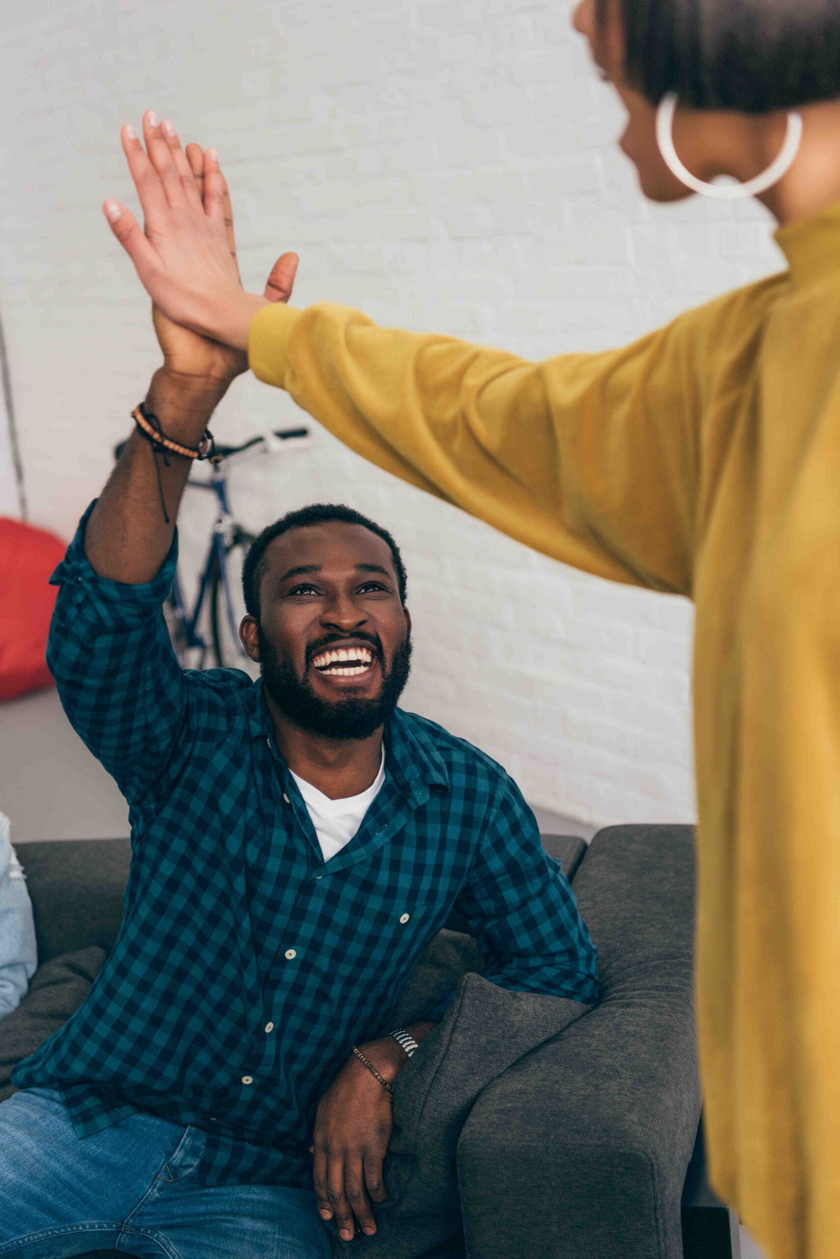 A man in a checkered shirt, relaxed on the couch, smiles as he high-fives a standing person in a mustard yellow sweater, capturing a moment of camaraderie akin to the supportive environment found in residential treatment settings.