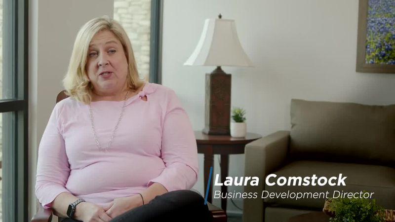 A woman in a light pink top sits in an armchair next to a side table and a lamp. The text on the image reads: Laura Comstock, Business Development Director.